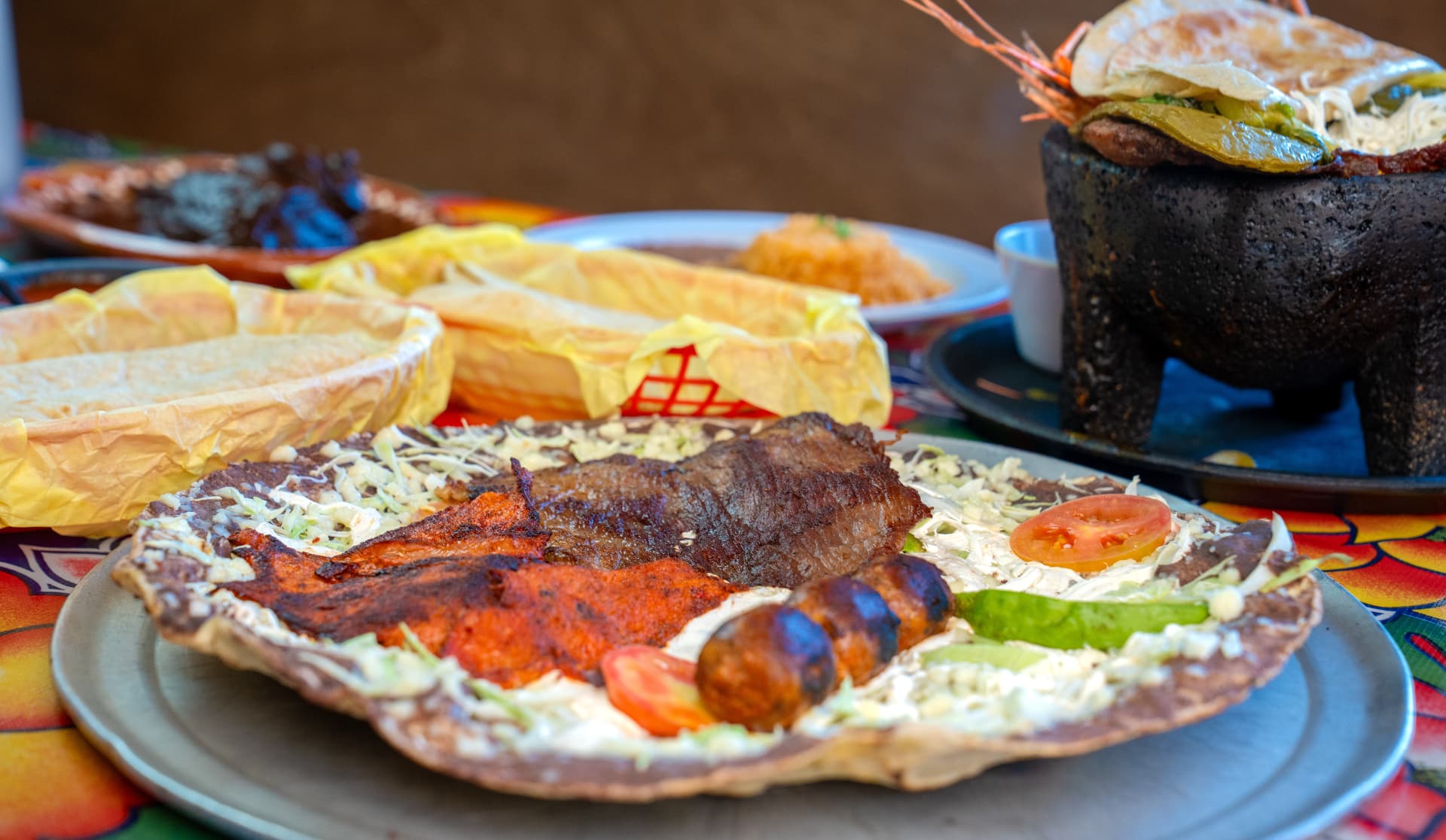A tlayuda topped with carne asada, cecina, and chorizo on a colorful Oaxacan tablecloth